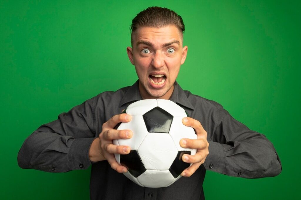 Hombre guapo joven en camisa gris con balón de fútbol gritando con expresión